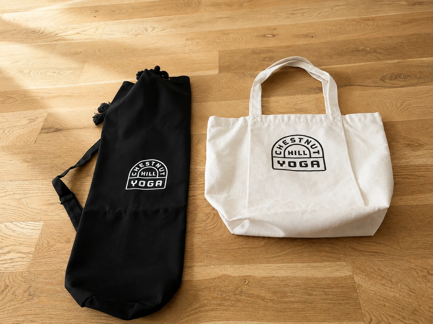 Retouched client photograph of a black mat bag and natural canvas tote on an oak floor, each printed with the Chestnut Hill Yoga crest.