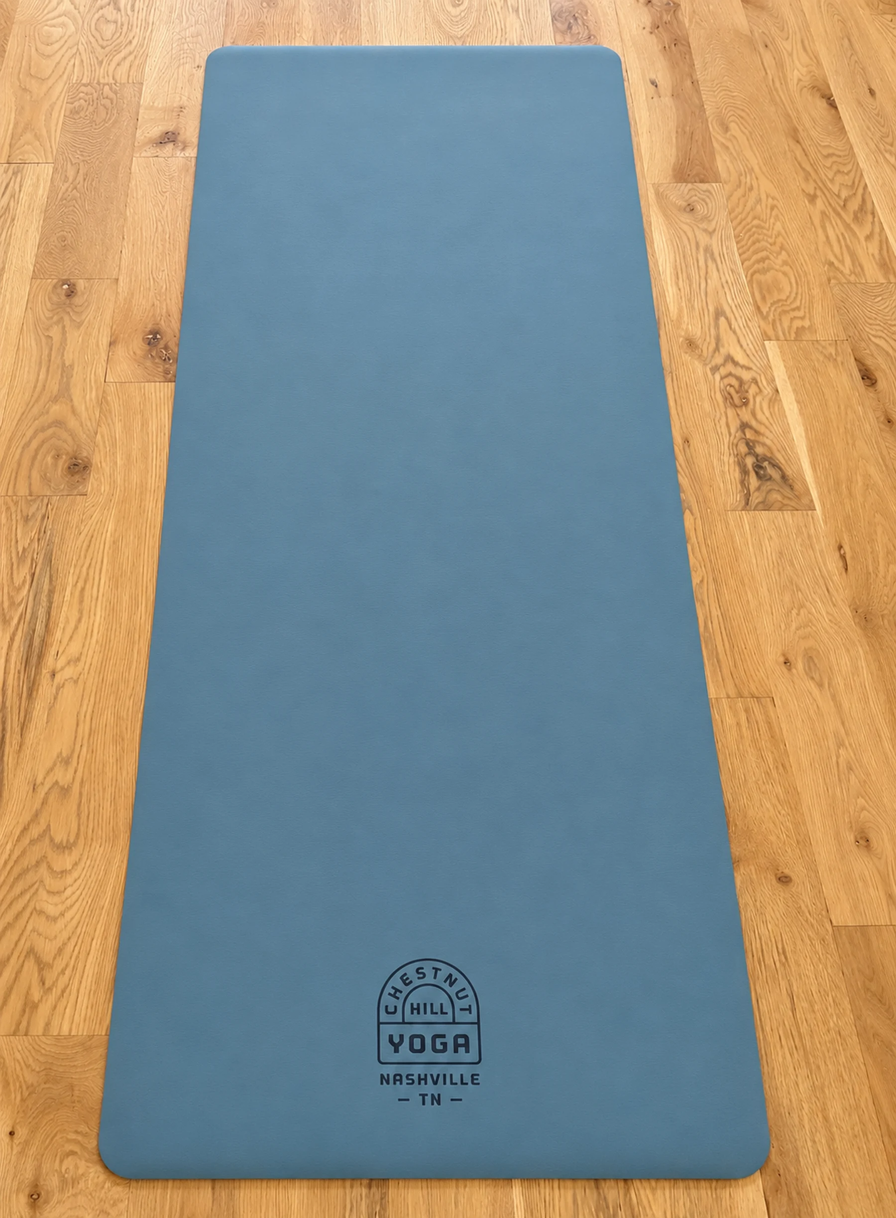 Full-length client photograph of a navy polyurethane Chestnut Hill Yoga mat with the studio crest, laid out on the studio’s oak floor.