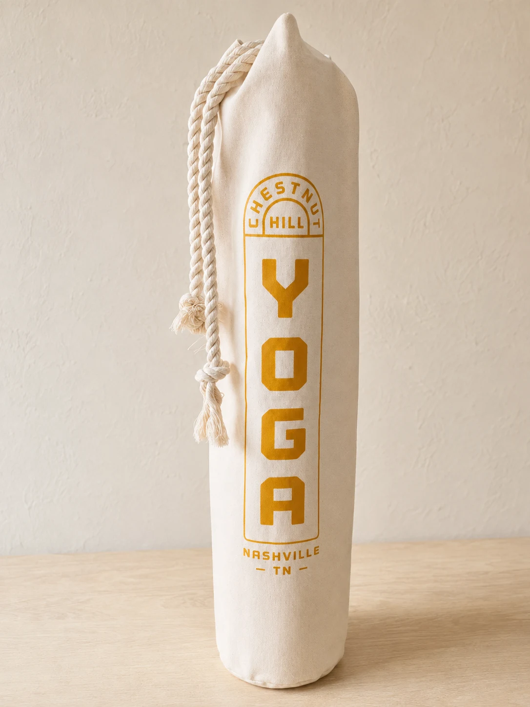 Natural canvas mat bag with thick rope drawstrings and an ochre Chestnut Hill Yoga Nashville TN print, photographed on a table.