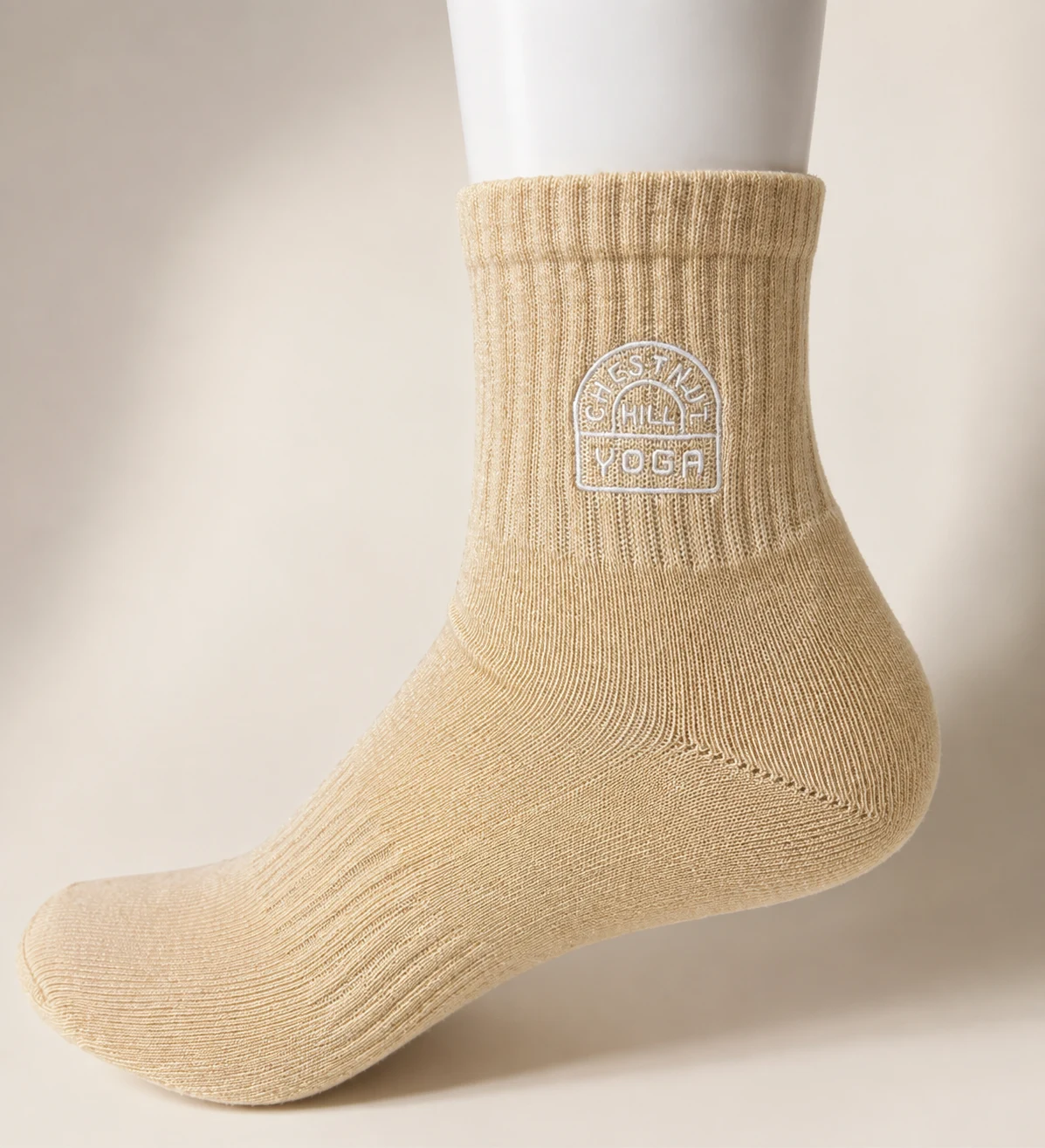 Retouched close-up of a Chestnut Hill Yoga crest embroidered in light thread on an oatmeal sock on a display form.